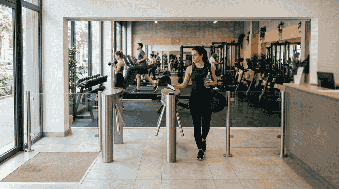 Vue depuis l'entrée d'une salle de fitness moderne où une femme utilise son smartphone pour passer un portique d'accès. En arrière-plan, plusieurs adhérents s'entraînent sur des tapis de course et des appareils de musculation.